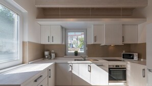 Een moderne keuken met witte kasten, een inbouwoven, een magnetron en een raam met zonwering.
