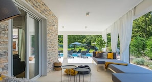 Un espace extérieur abrité, doté d’un mur en pierre, d’un canapé avec des coussins, d’une petite table et d’une piscine en arrière-plan.