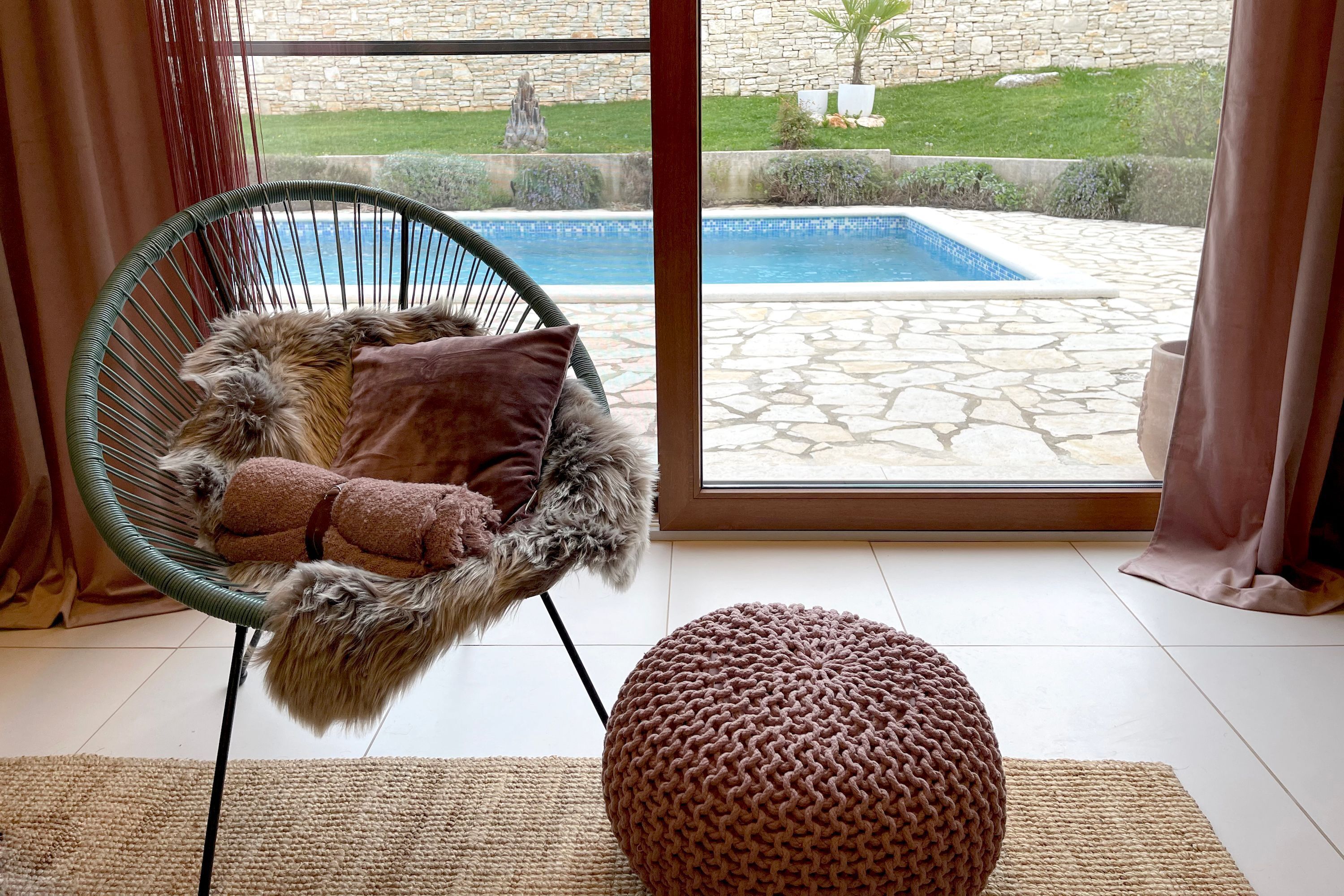 A cozy room with a wicker chair, a knitted ottoman, and a view of a swimming pool through a glass door.
