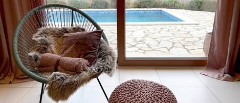A cozy room with a wicker chair, a knitted ottoman, and a view of a swimming pool through a glass door.