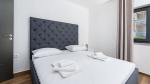 Una cama bien hecha con ropa de cama blanca y un cabecero gris, una mesita de noche con una lámpara, y una ventana con cortinas.