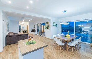 A modern living room with a sofa, a dining table with chairs, a kitchen area, and a view of a swimming pool. - OL JU in Pula With 3 Bedrooms and 1 Bathrooms (Pula)