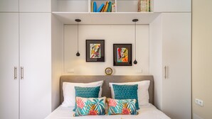 Ein Schlafzimmer mit einem Bett, zwei Kissen, einem Nachttisch, einem Regal mit Büchern und zwei gerahmten Kunstwerken an der Wand.