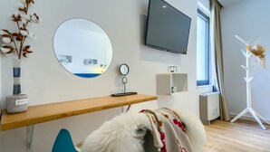 A modern hotel room with a wooden desk, a round mirror, a flat-screen TV, a window with curtains, a radiator, and a decorative hat on a stand. - Bellevue in Bast Ba ka Voda (Baska Voda)