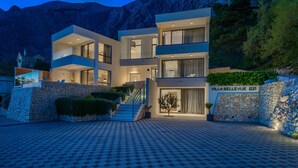Una villa moderna con fachada de piedra, varios balcones y un acceso empedrado.