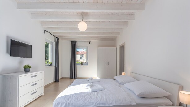 A modern bedroom with a large bed, a TV mounted on the wall, a bedside table with a lamp, and a view of the outside through a window. - Complex Kamenjak in Premantura (Medulin)
