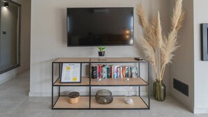 Un salón con un televisor de pantalla plana montado en la pared, una estantería con libros y objetos decorativos, una pequeña planta en maceta y plumas de pampa en un florero.