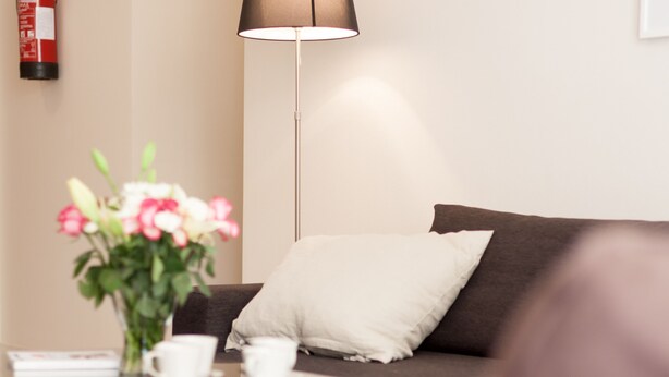 Una habitación moderna con un sofá marrón oscuro, un cojín blanco, una mesita con una taza y un platillo blancos, y una lámpara de pie.
