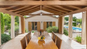 Eine überdachte Terrasse mit einem Holztisch, gedeckt für zwei Personen, einer Obstschale und einer Weinflasche.