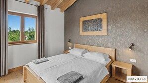 Ein Schlafzimmer mit Holzboden, einem Bett mit weißer Bettwäsche, zwei Nachttischen mit Lampen, einem Fenster mit Vorhängen und einer Wand mit strukturierter Oberfläche und einem gerahmten Bild.