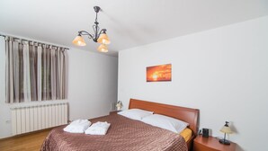 A neatly made bed with a brown bedspread, a wooden headboard, and white pillows. There is a nightstand with a lamp and a small table with a radio. A window with curtains and a radiator are visible. - Marina Opatija in Opatija (Opatija)