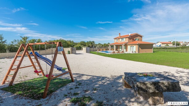 A playground with a swing set and slide, a stone bench, and a house with a red roof in the background.