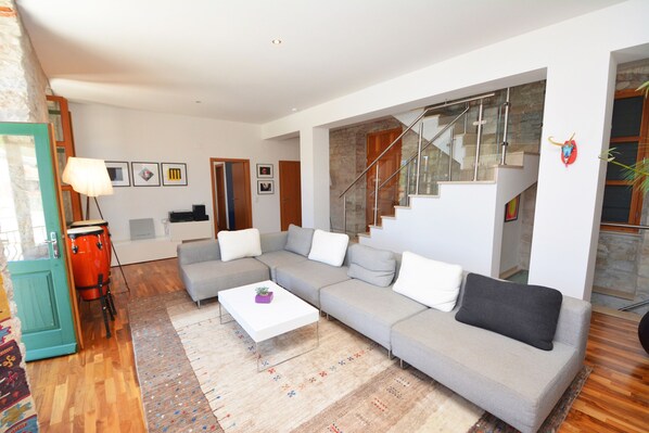 Un salon moderne avec un canapé d’angle gris, une table basse blanche et un tapis à motifs. Un escalier avec des rampes en verre mène à l’étage supérieur.