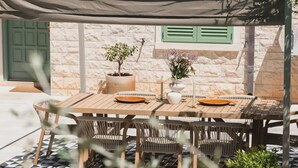 Un espace extérieur abrité, aménagé pour prendre les repas, avec une table et des chaises, un pot de fleurs et une vue sur un bâtiment dont le toit est recouvert de tuiles.