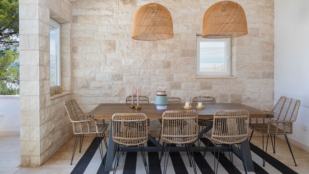 Un coin repas agrémenté d’un mur en pierre, de chaises en osier, d’une table en bois et de lampes en osier suspendues.