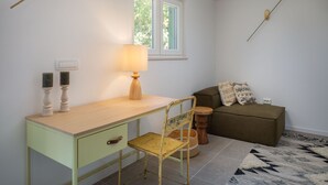 Une pièce comprenant un bureau, une chaise, une lampe, une fenêtre et un canapé avec des coussins.