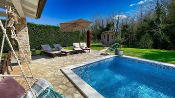 A swimming pool with lounge chairs and a hanging chair, surrounded by a stone patio and a small house in the background.