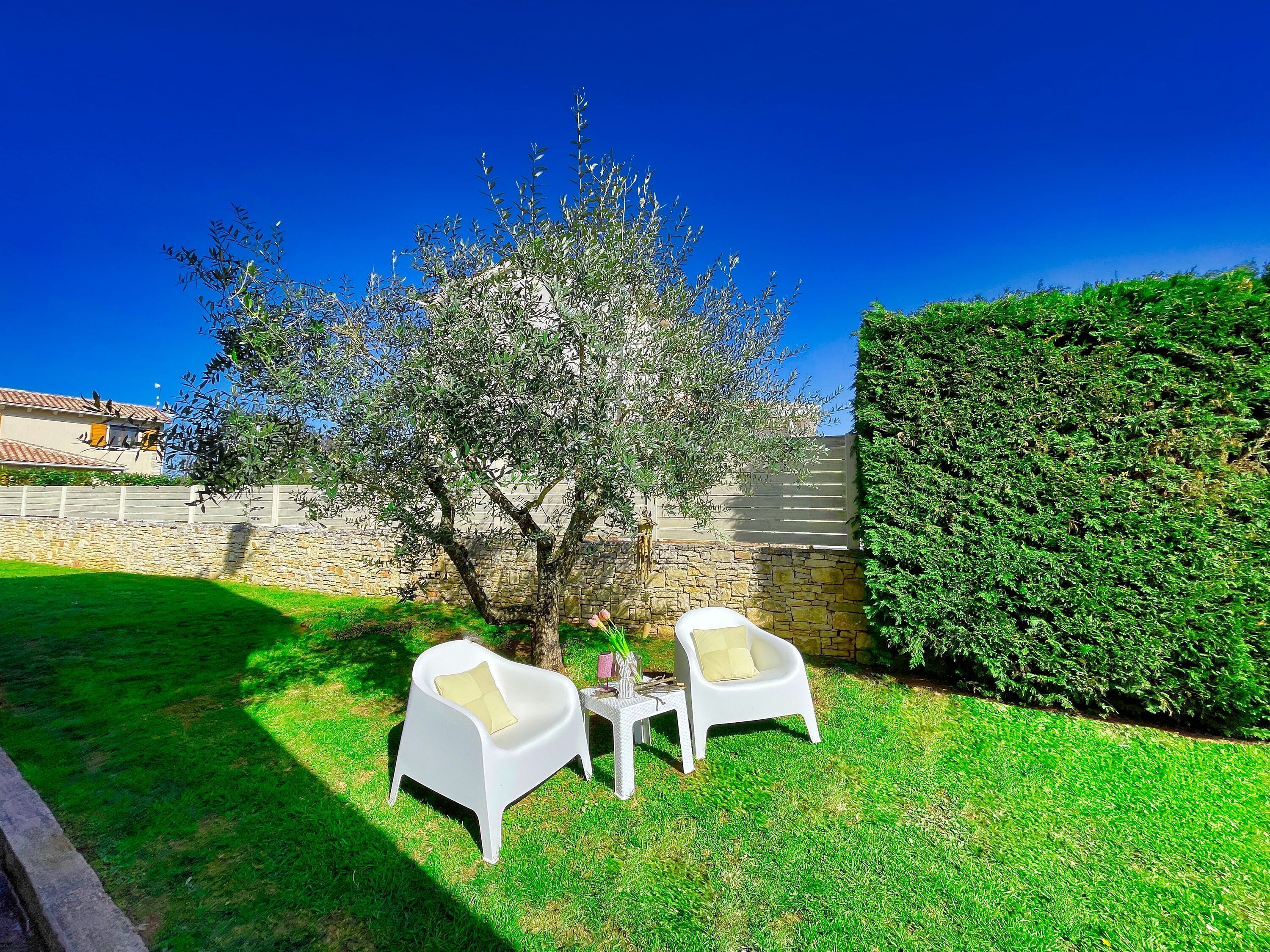 A patio with two white chairs and a table, surrounded by a well-maintained lawn and a stone wall.