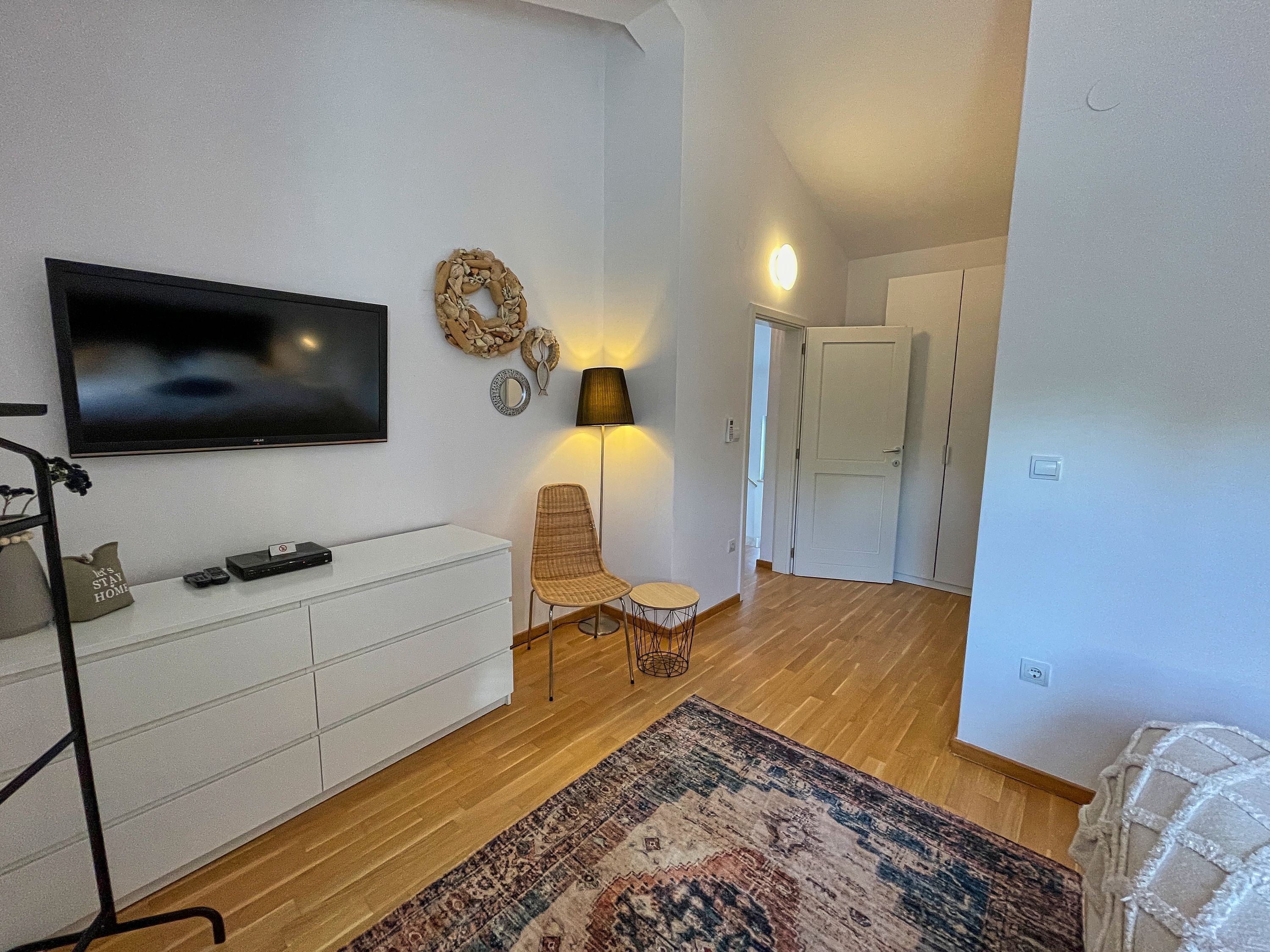 A modern living room with a flat-screen TV, a white cabinet, a wicker chair, a small round table, a patterned rug, and a hallway leading to a door.