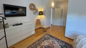 Un saló modern amb un televisor de pantalla plana, un moble blanc, una cadira de vimet, una taula petita rodona, una catifes amb dibuixos i un passadís que condueix a una porta.