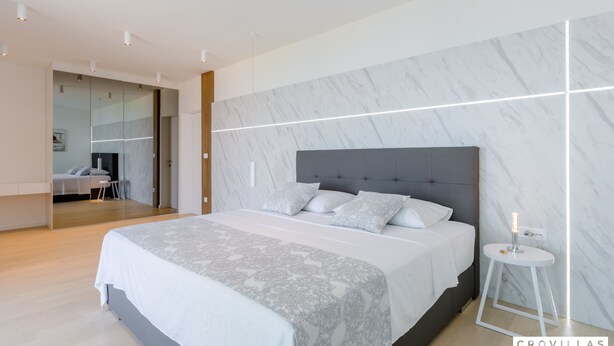Un dormitorio moderno con una cama grande, paredes con diseño de mármol y una mesita de noche pequeña con una lámpara.
