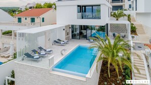 Una villa moderna con piscina en la azotea, sillas de descanso y un patio con cerramiento de vidrio.