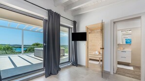 Une chambre d’hôtel moderne dotée d’une grande porte-fenêtre coulissante donnant sur un balcon, d’un téléviseur à écran plat fixé au mur et d’une porte en bois menant à la salle de bain.