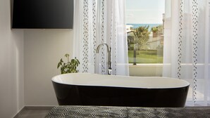 Un baño moderno con una bañera independiente, un televisor montado y vistas al exterior a través de cortinas translúcidas.