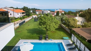 Una piscina con sillones de playa y sombrillas, rodeada por un césped cuidado y vista al mar.