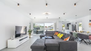 A modern living room with a grey sectional sofa, a flat-screen TV, and a dining area with a table and chairs. - Elite 1 in Pula With 4 Bedrooms and 4 Bathrooms (Pula)