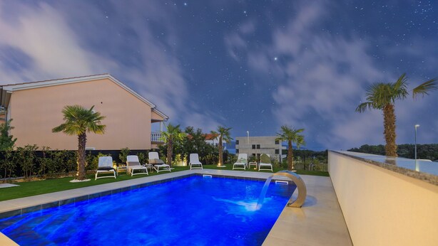 Piscina com espreguiçadeiras, palmeiras e um edifício ao fundo sob um céu estrelado.