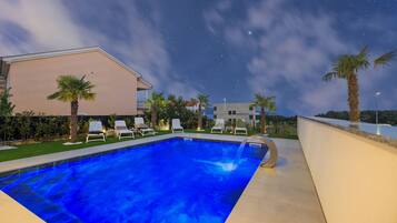 A swimming pool with lounge chairs, palm trees, and a building in the background under a starry night sky.
