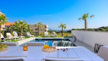 A poolside area with lounge chairs, a dining table with a basket of fruit and a drink, and a view of the pool and surrounding landscape.