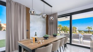Uma zona de refeições moderna com uma mesa de madeira, seis cadeiras e uma janela ampla com vista para uma piscina e palmeiras.