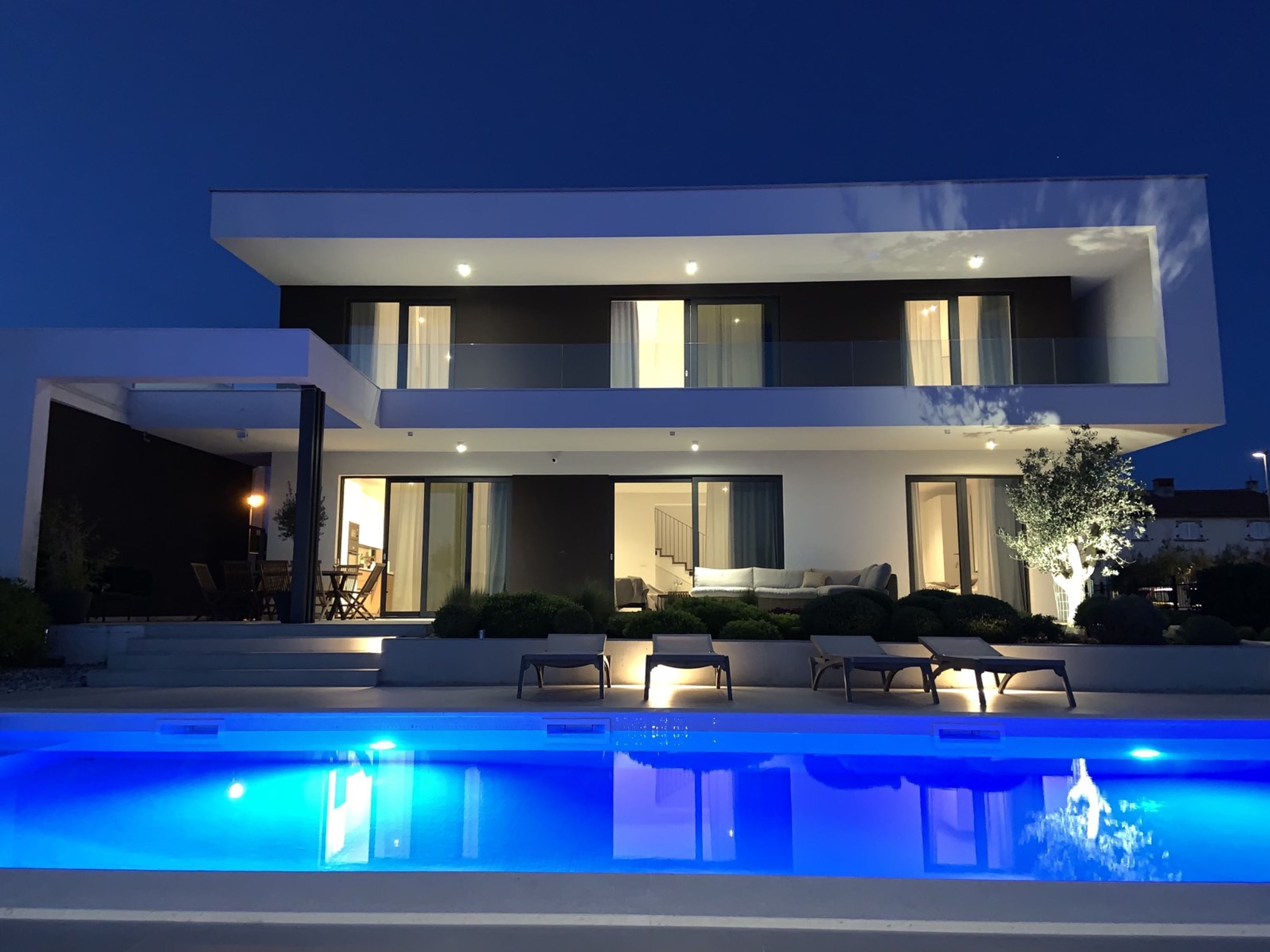 Una casa moderna de dos pisos con piscina, mobiliario exterior y una entrada bien iluminada.