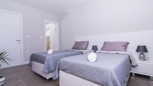 A twin bedroom with two beds, each with a pillow and a rolled towel. There is a nightstand with a lamp and a small table with a decorative item. - Kastel One in K Kambelovac (Kastela)