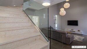A modern interior with a staircase, glass railings, a grey sofa, a flat-screen TV, and a console table. - Kastel One in K Kambelovac (Kastela)