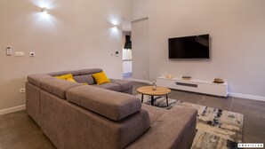 A modern living room with a sectional sofa, a flat-screen TV mounted on the wall, and a small side table. - Kastel One in K Kambelovac (Kastela)