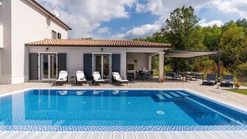 A swimming pool with lounge chairs, a covered patio, and a house with a tiled roof.