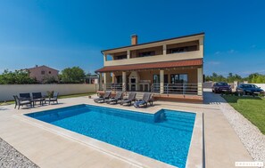 Una casa de dos pisos con piscina, mobiliario exterior y cochera.
