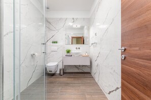 Un baño moderno con paredes de mármol, suelo de madera, cabina de ducha de vidrio, lavamanos blanco y espejo sobre el lavamanos.