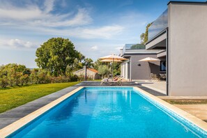 Una piscina moderna con sillones de descanso y sombrillas, rodeada por un césped cuidado y un edificio de piedra.