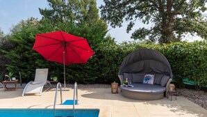 Ein Poolbereich mit rotem Sonnenschirm, Liegestuhl, runder Sitzecke und kleinem Tisch mit Kerze und Glas.