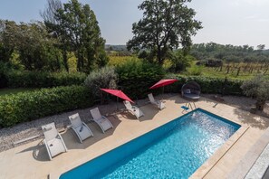 Ein rechteckiger Swimmingpool mit gefliestem Rand, umgeben von Liegestühlen und roten Sonnenschirmen.