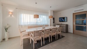 A modern dining area with a large table and chairs, a TV mounted on the wall, and a wooden door with glass panels. - Dalmacija VI in Suko an (Sukosan)