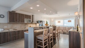 A modern kitchen with wooden cabinets, a central island with bar stools, a dining area with a table and chairs, and recessed ceiling lighting. - Dalmacija VI in Suko an (Sukosan)