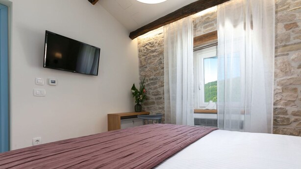 A bedroom with a bed, a TV mounted on the wall, a stone wall, a window with curtains, and a small desk with a chair. - Flego in Motovun With 2 Bedrooms and 1 Bathrooms (Motovun)