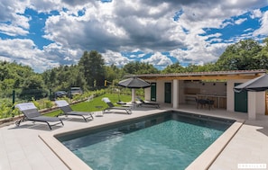 Une piscine avec des chaises longues et des parasols, une cuisine extérieure couverte et une vue sur un paysage verdoyant luxuriant.