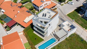 Une maison moderne à deux étages, dotée d’une piscine, d’un patio couvert et d’un toit recouvert de tuiles rouges.