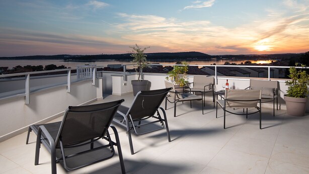 Une terrasse sur le toit, aménagée avec des espaces de détente en plein air, des plantes en pot et offrant une vue sur le coucher de soleil au-dessus d’une étendue d’eau.